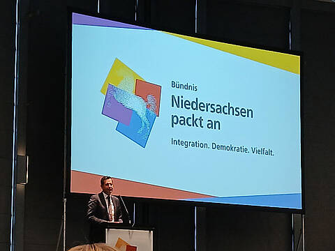 Ministerpräsident Olaf Lies hält eine Rede an einem Podium vor einer großen Leinwand mit der Aufschrift "Bündnis Niedersachsen packt an. Integration. Demokratie. Vielfalt". Dies bezieht sich auf das Bündnis "Niedersachsen packt an", eine Initiative f